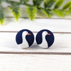 May include: A pair of blue fabric button earrings with white goose designs. The geese have orange beaks.
