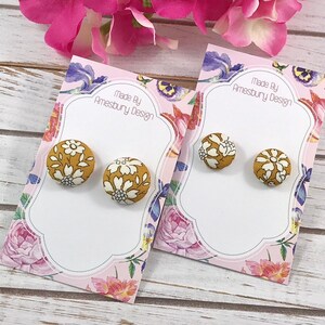 May include: A pair of mustard yellow button earrings with a white floral pattern. The earrings are displayed on a card with the text "Made By Amesbury Design".