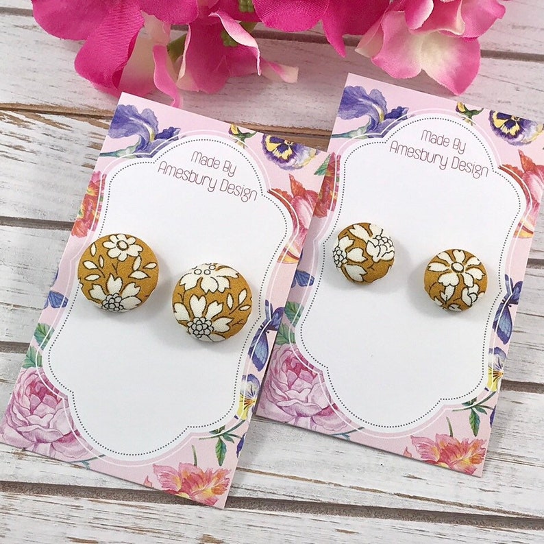 May include: A pair of mustard yellow button earrings with a white floral pattern. The earrings are displayed on a card with the text "Made By Amesbury Design".