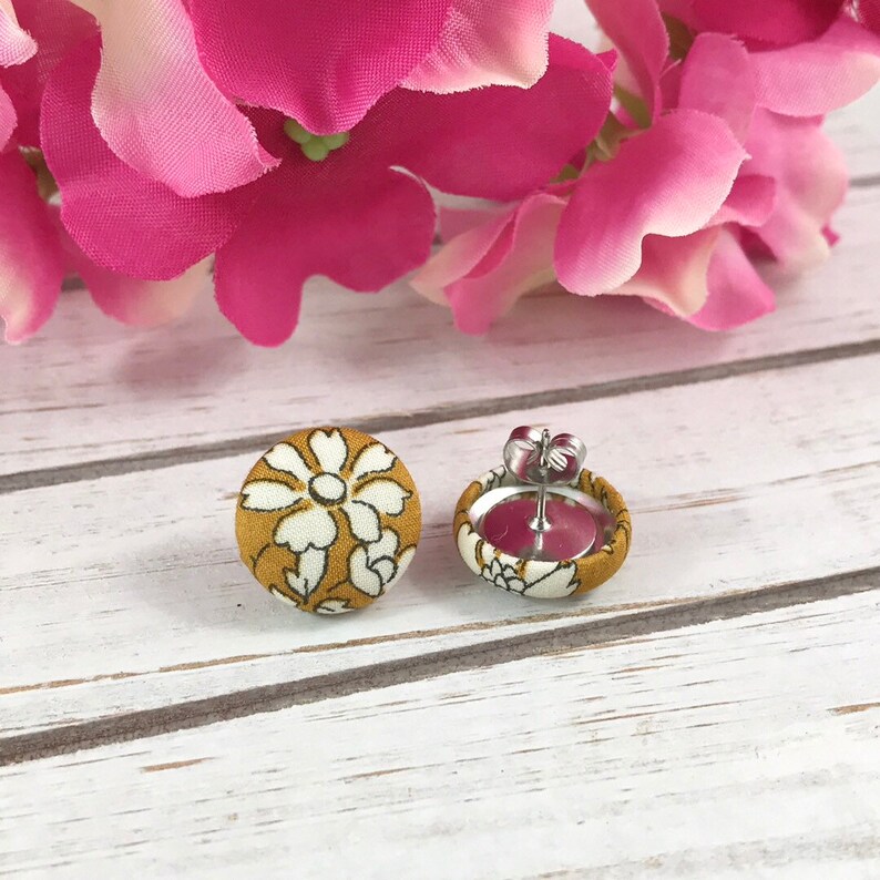 May include: A pair of mustard yellow fabric earrings with a white floral pattern. The earrings are stud style with a silver post and butterfly backing.
