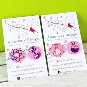 May include: Two sets of pink button earrings with science-themed designs. The first set features an atom and a DNA double helix. The second set features a beaker and a flask. The earrings are handmade and come with a card that says "Amesbury Design Handmade Jewellery & Accessories" and includes contact information for the seller.