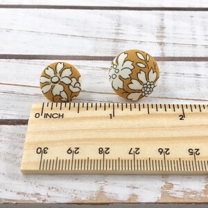May include: Two fabric buttons with a floral pattern in white and mustard yellow. The buttons are shown next to a ruler, measuring approximately 3/8 inch in diameter.