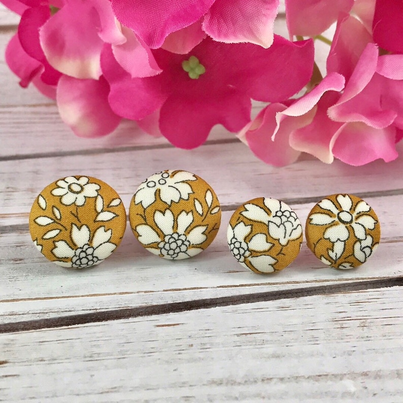 May include: Four round fabric buttons with a floral pattern in white on a mustard yellow background. The buttons are arranged in a row on a white wooden surface.