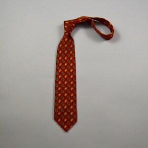 May include: A red necktie featuring a repeating pattern of small, gold-toned paisley shapes. The necktie is tied in a knot at the top, with the narrow end looped around to the side, displaying the consistent fabric print throughout the length.