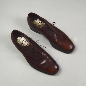 May include: A pair of dark brown leather dress shoes with laces. The shoes have a classic design with decorative perforations on the toe and sides. The soles are dark brown, and the shoes appear to be new.