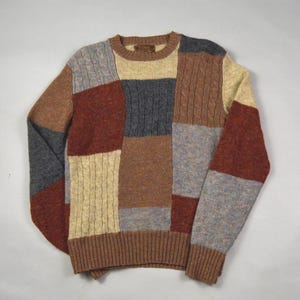May include: A multi-coloured patchwork sweater with a crew neck and ribbed cuffs and hem. The sweater's design incorporates various shades of brown, grey, red, and beige, with cable knit and textured patterns.