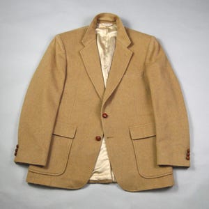 Könnte beinhalten: Ein beigefarbener Blazer mit klassischem Design. Die Jacke hat ein fallendes Revers, zwei Vordertaschen und braune Knöpfe. Das Innenfutter ist hellgoldfarben. Diese maßgeschneiderte Jacke eignet sich für verschiedene Anlässe.