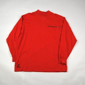 May include: Bright red long-sleeve shirt with a mock neck and a small zippered pocket on the chest. The shirt has a small logo and the text "RL-83" printed in black near the bottom hem. The sleeves have ribbed cuffs.
