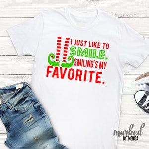 May include: White t-shirt with the text "I just like to smile. Smiling's my favourite" in red and green. The design includes elf legs and the word "smile" in green polka dots. A pair of blue jeans and trainers are in the background.