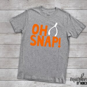 May include: A heather gray t-shirt with the words "OH SNAP!" in bright orange, and a white wishbone graphic. The shirt is made of a soft material and is a casual wear item.