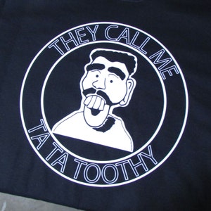May include: Black t-shirt with a white circle graphic. The graphic features a cartoon character with a tongue sticking out and the text "They Call Me Tata Tootty".