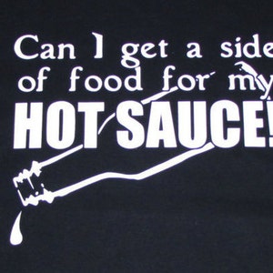 May include: Black t-shirt with white text that reads "Can I get a side of food for my HOT SAUCE!" with a white silhouette of a hot sauce bottle pouring out sauce.