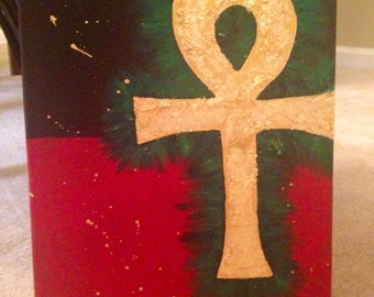 May include: A gold Ankh symbol painted on a canvas with a black, green, and red background. The symbol is painted with gold leaf and has a textured look. The canvas is signed "D. Rosenthal" in the bottom left corner.