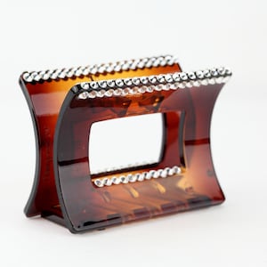 May include: A brown tortoiseshell hair claw clip with a rectangular opening in the center. The top and bottom edges are embellished with a row of small, clear rhinestones. The clip has a curved, arched design.
