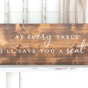 May include: A brown wooden sign with white text that reads "At every table I'll save you a seat".