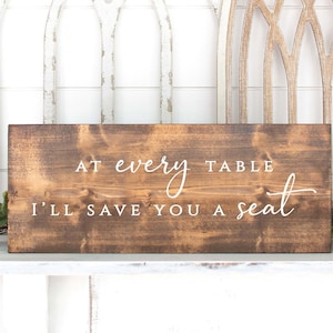 May include: A brown wooden sign with white text that reads "At every table I'll save you a seat".