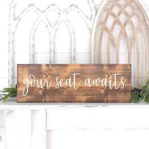 May include: A brown wooden sign with white lettering that reads "your seat awaits".