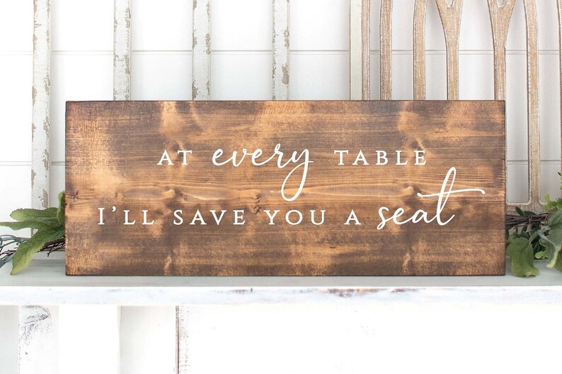 May include: A brown wooden sign with white text that reads "At every table I'll save you a seat".