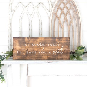 May include: A brown wooden sign with white text that reads "At every table I'll save you a seat".