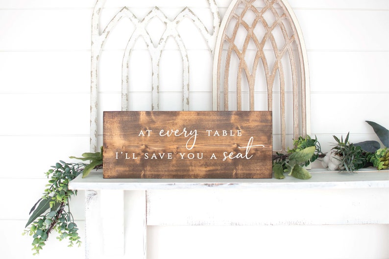 May include: A brown wooden sign with white text that reads "At every table I'll save you a seat".