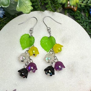 Peut inclure: Une paire de boucles d'oreilles pendantes avec des feuilles vertes et une cascade de perles de fleurs violettes, noires, blanches et jaunes. Les boucles d'oreilles sont présentées sur une surface circulaire blanche.