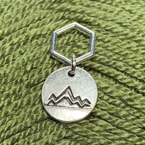 May include: A silver knitting stitch marker with a hexagonal ring and a round disc featuring a mountain range design.