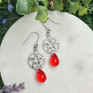 May include: Silver earrings with a pentagram charm and red glass teardrop dangles. The earrings are displayed on a white circular surface with a floral background.