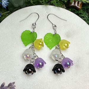 May include: A pair of dangle earrings featuring a cluster of colorful flowers and green leaves. The earrings have a silver hook and are set against a white marble surface, surrounded by greenery and flowers.