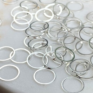 May include: A large quantity of silver jump rings, scattered on a white surface and a wooden surface. The jump rings are small and round, with a smooth, polished finish.