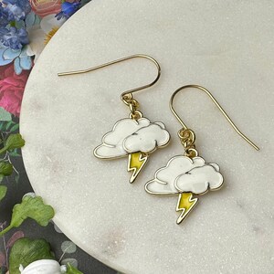 Peut inclure: Une paire de boucles d'oreilles pendantes dorées avec des breloques en forme de nuage blanc et des accents de foudre jaune. Les boucles d'oreilles sont présentées sur une surface en marbre blanc.