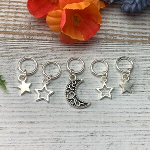 May include: Five silver stitch markers with star and crescent moon charms. The charms are on a white wooden background.