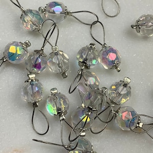 May include: A group of 15 clear iridescent glass stitch markers with silver metal loops. The markers are scattered on a white surface.