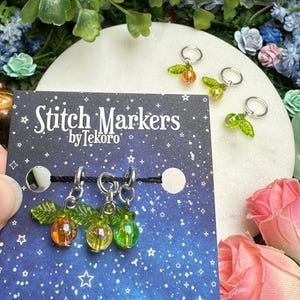 May include: A set of three stitch markers with orange and green bead charms, each with a leaf accent. The markers are attached to a black string and displayed on a card with the text "Stitch Markers by Tekoro" against a starry background.