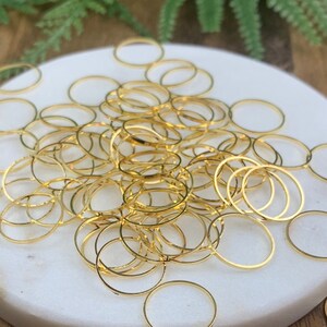 May include: A selection of small, gold-coloured metal rings scattered on a white marble surface and a wooden table. The rings are thin and circular, with a shiny finish. Some rings are grouped together, while others are isolated.