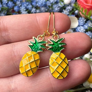 May include: A pair of gold pineapple earrings with green tops and yellow bodies. The earrings are on a hand against a background of flowers.