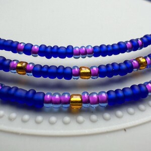 May include: A triple-strand beaded necklace with blue, purple, and gold beads. The necklace is made of small, round beads and is layered in a simple design.