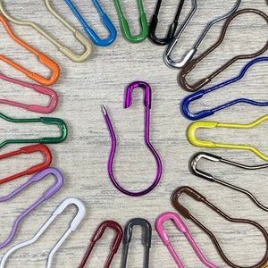 May include: A collection of 20 colorful safety pins in various sizes and shapes, arranged in a circle on a wooden surface.