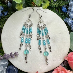 May include: A pair of silver dangle earrings with turquoise and light blue beads. The earrings have a teardrop shape and are accented with silver chains.