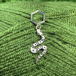 May include: A silver snake stitch marker with a hexagonal ring. The marker is on a green yarn ball.