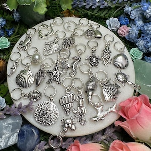 May include: A collection of silver-toned metal charms featuring various marine life and ocean-themed shapes, including seashells, a starfish, a sea turtle, a seahorse, a shark, and an octopus, each attached to a small circular metal jump ring.