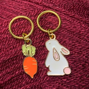 May include: Two gold stitch markers with charms. One charm is a white bunny with pink ears and nose. The other charm is an orange carrot with green leaves.