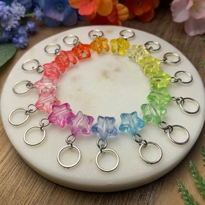 May include: A collection of colorful star-shaped stitch markers in a rainbow gradient. Each marker has a silver-colored ring attached. The markers are arranged in a circular pattern on a white marble surface.