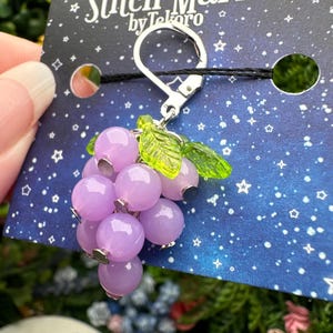 May include: A grape-shaped stitch marker featuring translucent light purple spherical beads and two bright green leaf-shaped accents attached to a silver-toned metal lever-back clasp, presented on a dark blue card labeled Stitch Markers by Tekoro.