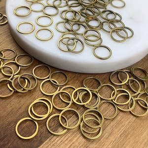 May include: A large quantity of gold-colored metal jump rings. The rings are scattered on a white surface and a wooden surface.