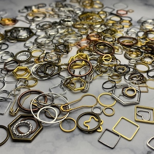 May include: A large assortment of metal jump rings, charms, and other jewelry findings in gold, silver, and bronze tones. The findings are scattered on a white surface.