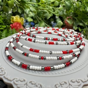 May include: A set of five beaded waist beads in white, red, and silver. The beads are strung on a thin cord and are arranged in a repeating pattern.