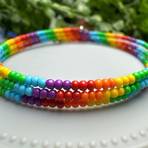 May include: A multi-colored beaded bracelet with a rainbow pattern. The bracelet is made of small, round beads in shades of red, orange, yellow, green, blue, indigo, and violet.