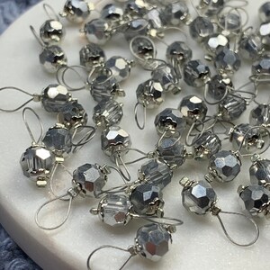 May include: A close-up of a pile of silver-toned metal stitch markers with clear glass beads. The beads are faceted and have a shiny finish. The stitch markers are used for marking rows in knitting and crocheting.