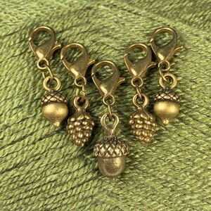 May include: Set of five gold-toned acorn stitch markers with a textured finish. Each marker features a different acorn design.