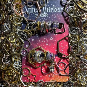 May include: A close-up of a card with the text "Stitch Markers by Tekoro" on a red background with white stars. Two sets of stitch markers are displayed on the card, one set is silver and the other is gold. The stitch markers are arranged in a circle shape and are surrounded by a variety of metal rings and hoops.